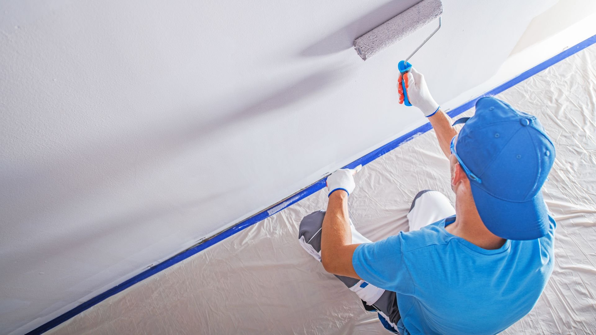 Painter in blue attire using roller to paint wall with protective drop cloth