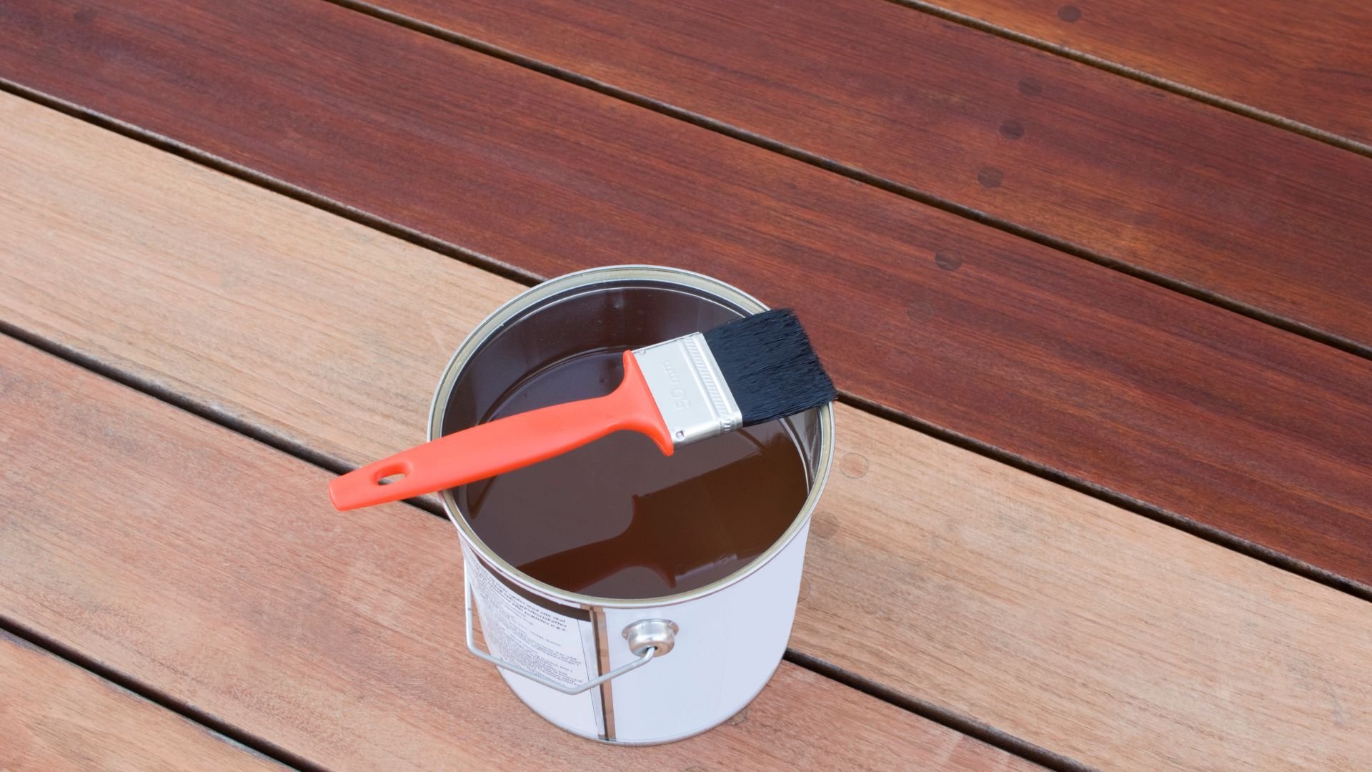 Red paintbrush resting on an open can of wood stain on wooden deck